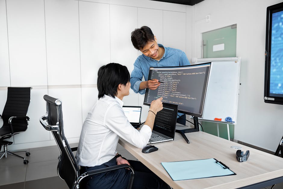Two young professionals collaborating at a desk with laptops and monitors in a modern office setting.