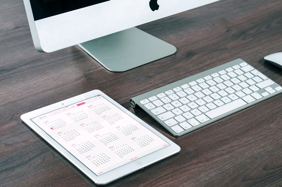 A sleek and modern office desk setup featuring an iMac, iPad with calendar, keyboard, and mouse.