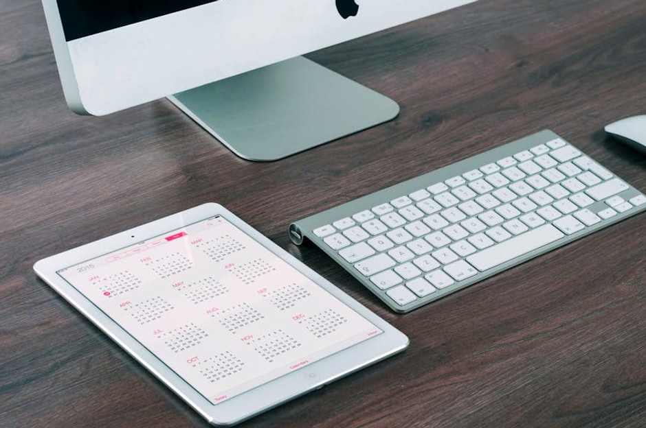 A sleek and modern office desk setup featuring an iMac, iPad with calendar, keyboard, and mouse.