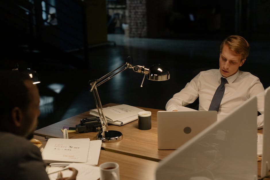 Two professionals working late in an office, reviewing documents under a desk lamp.