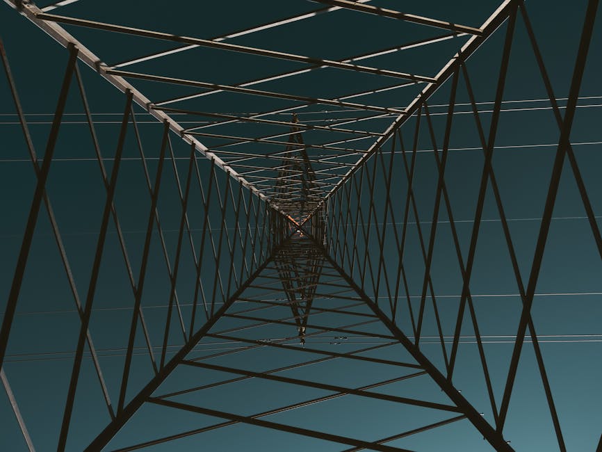 Abstract view looking up inside a transmission tower under a clear blue sky, creating a symmetrical pattern.