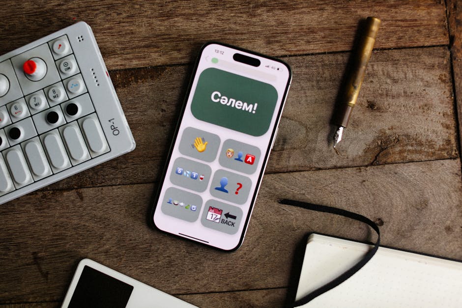 Smartphone displaying an app next to an OP-1 synthesizer, pen, and notebook on a wooden surface.