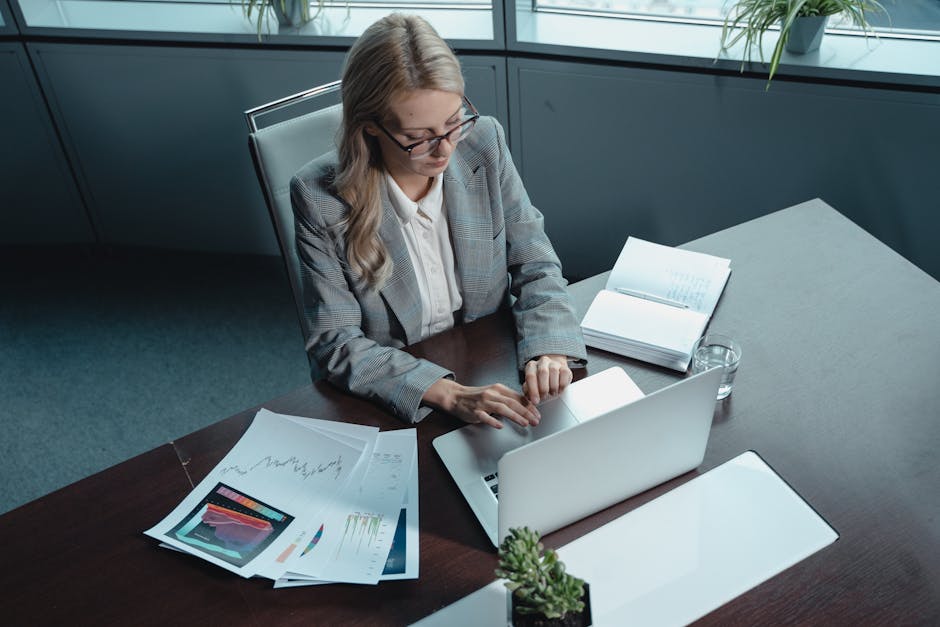 Blonde woman in a blazer working on a laptop in a modern office with documents.
