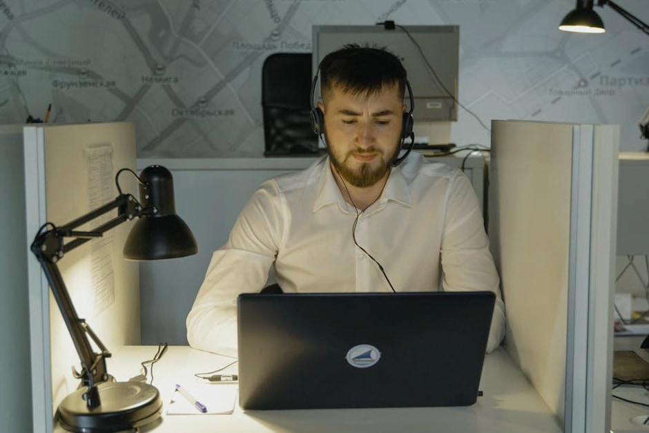 A bearded call center agent wearing headphones, focused on his laptop at work in a modern office.
